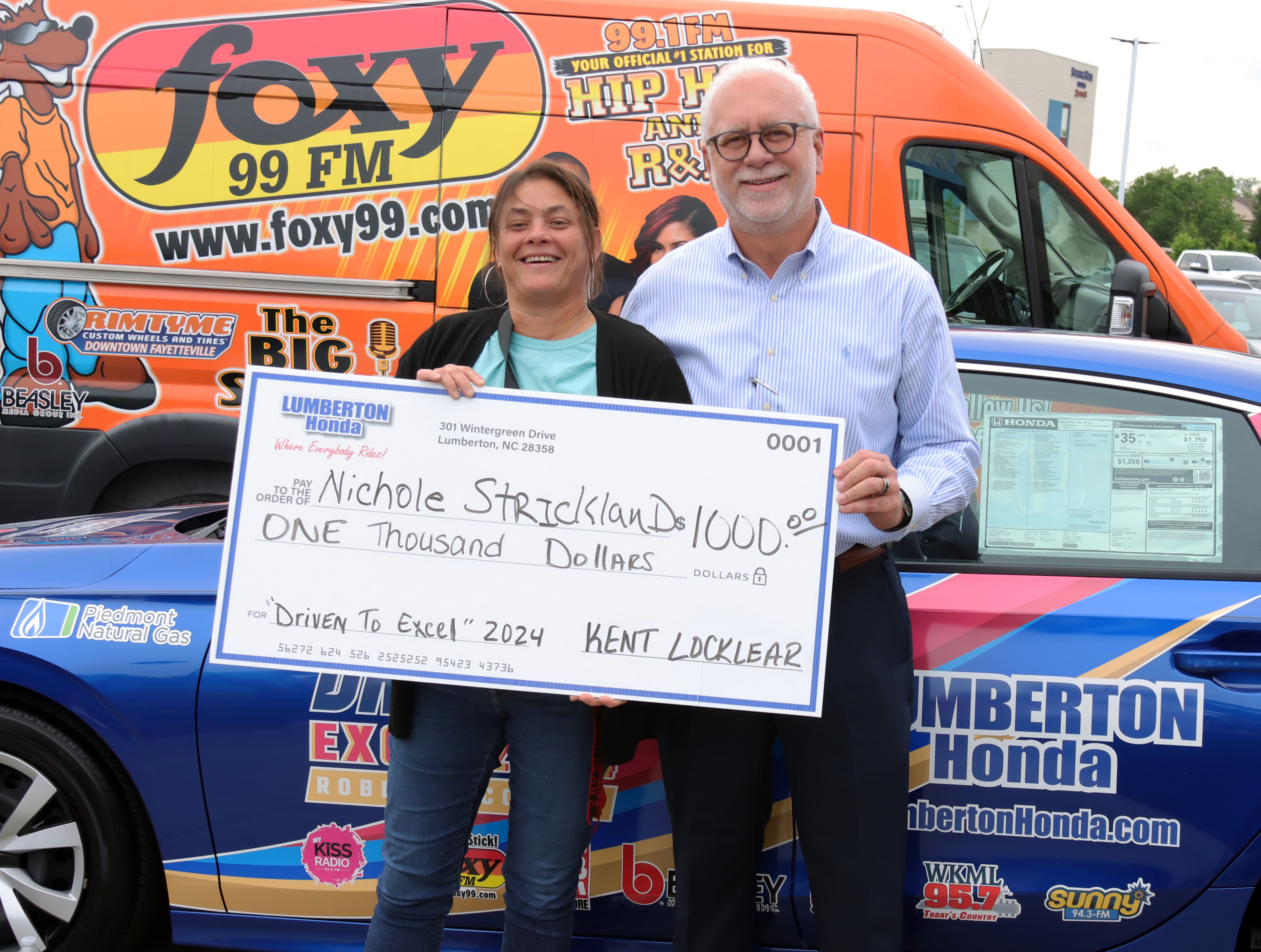 Two people holding a large check for $1000 in front of a Foxy 99 promotional van