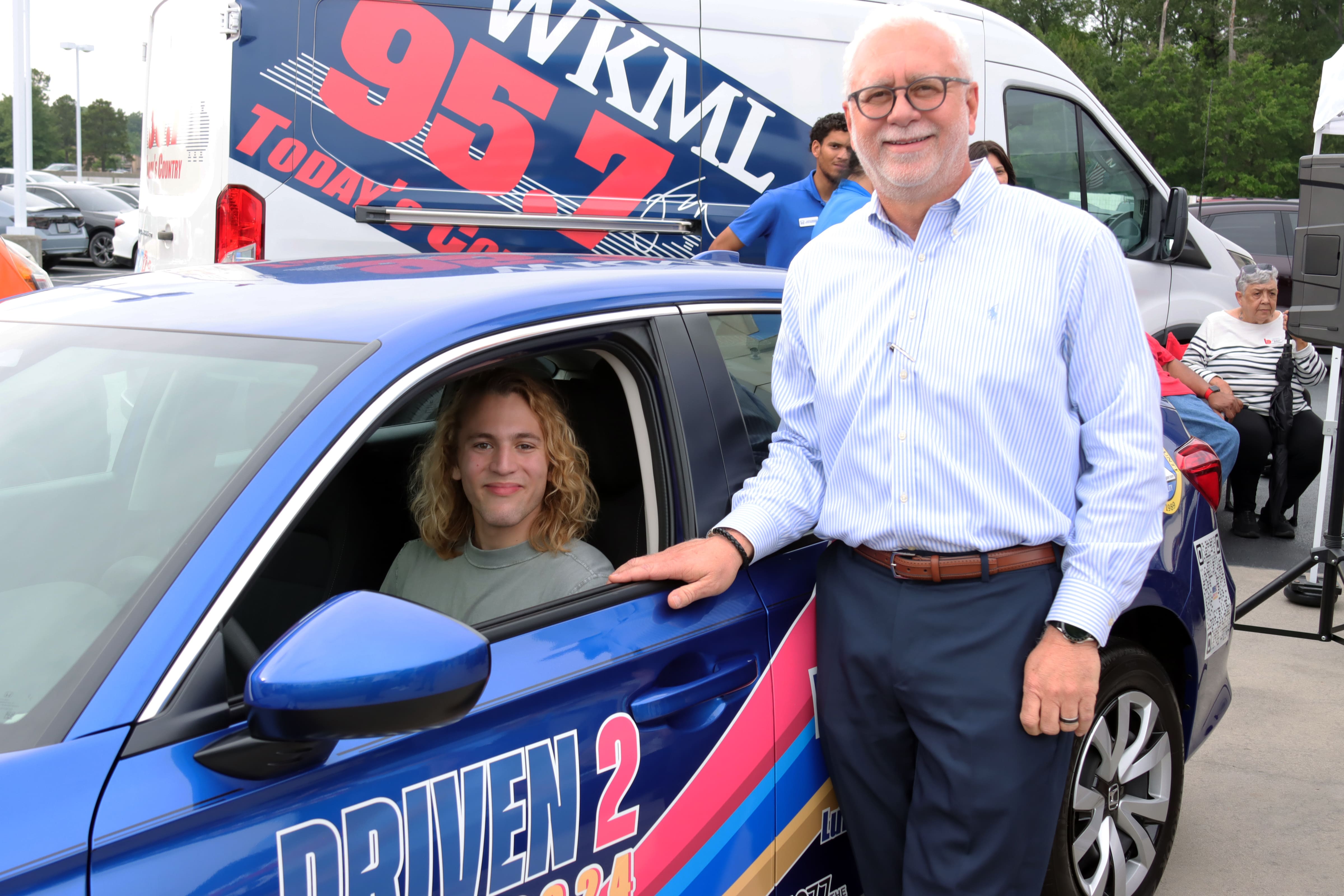 Man standing next to a car with another person sitting inside, in front of a WKML 95.7 promotional van